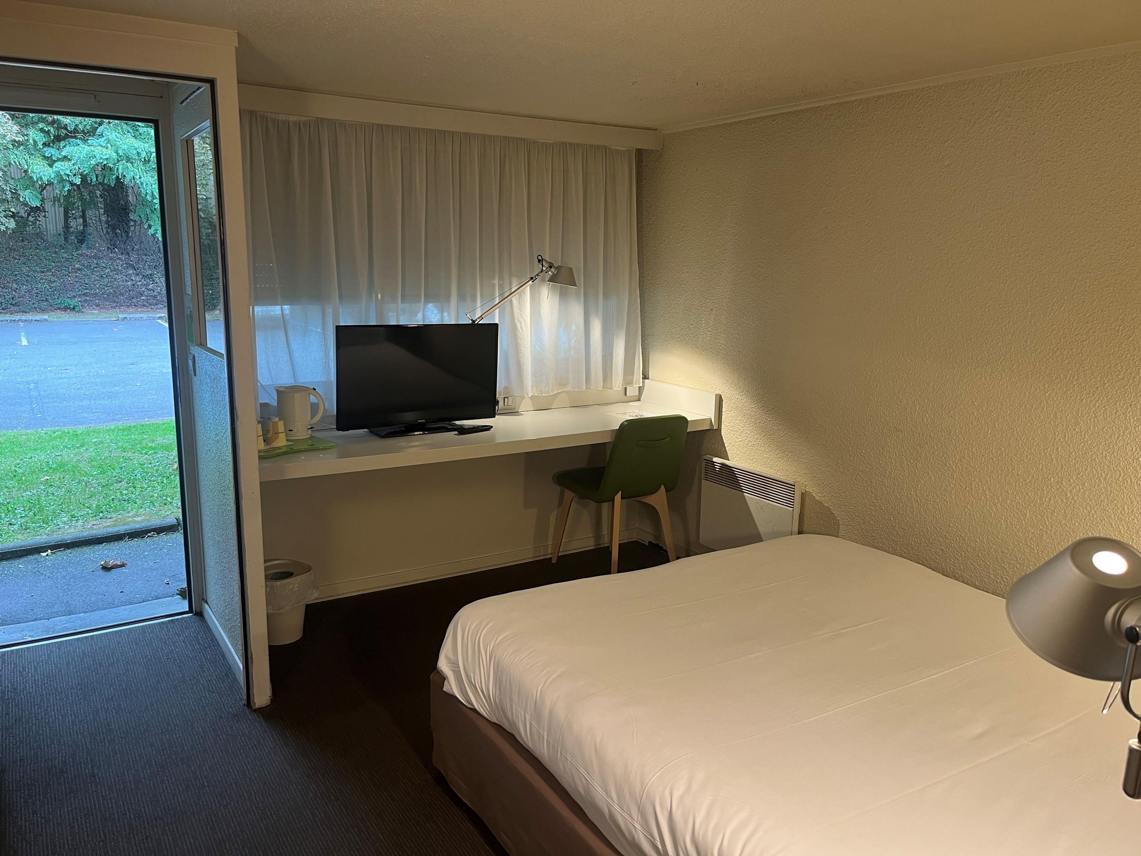 A hotel room with a bed, a desk with a computer, a chair, and a window with curtains.