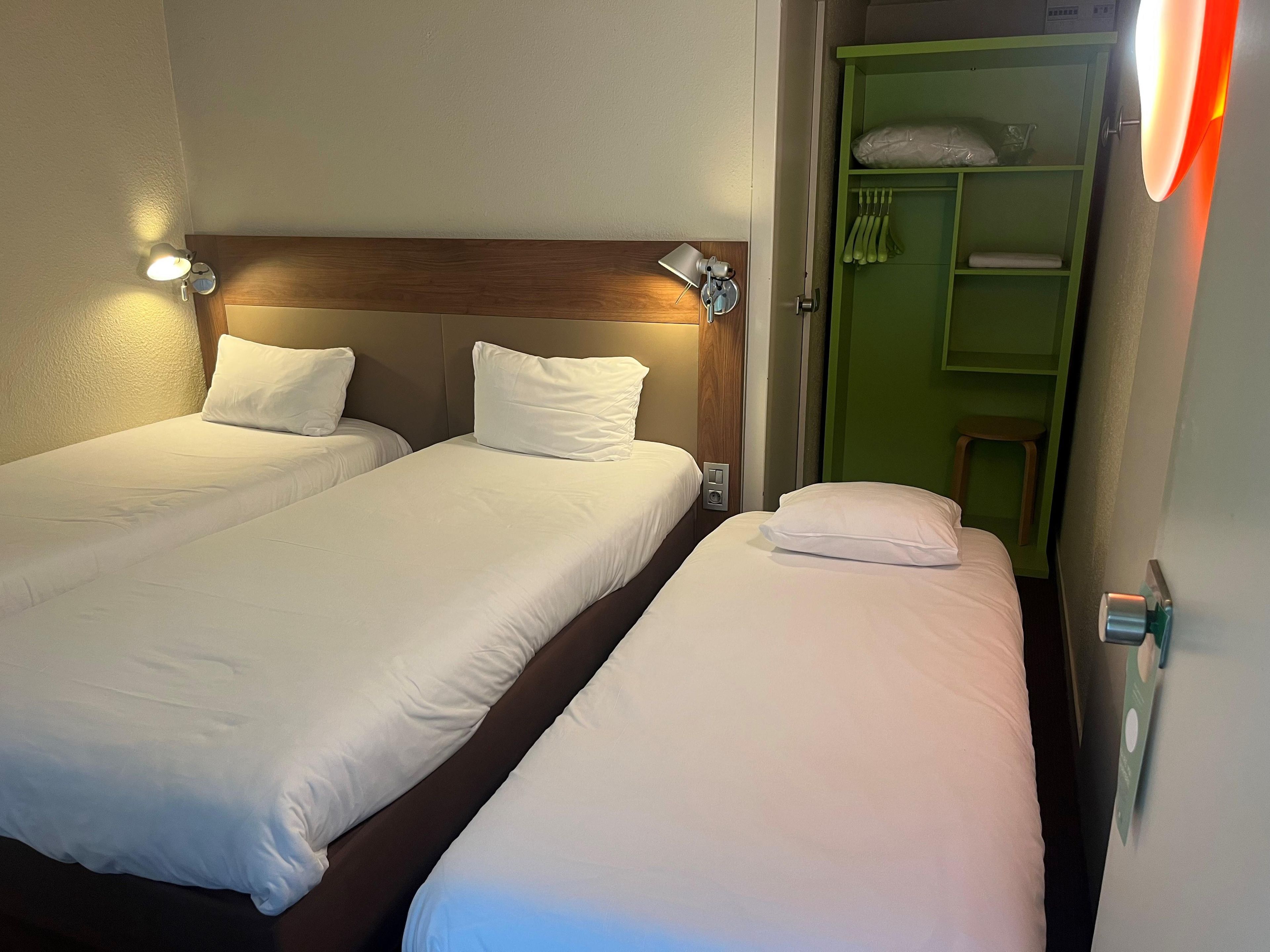 Dos camas individuales con sábanas blancas, un cabecero de madera y un mueble de almacenamiento verde con prendas colgadas.