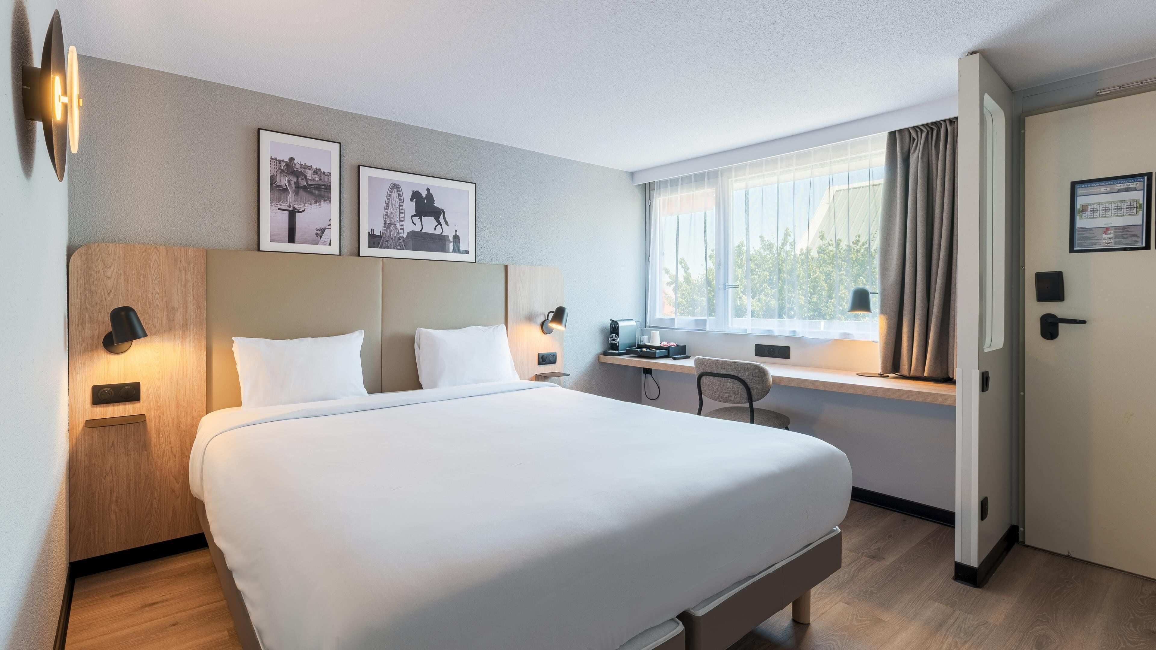 A hotel room with a large bed, a desk, a chair, two wall-mounted lamps, and two framed pictures on the wall.