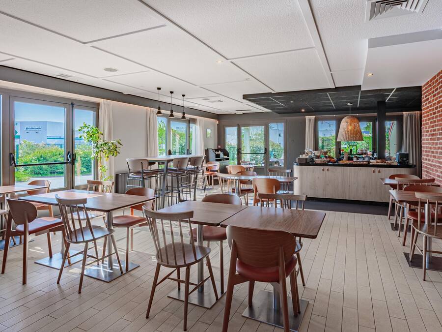 A modern dining area with multiple tables and chairs, large windows offering views of the outdoors, and a kitchen area with a brick wall.