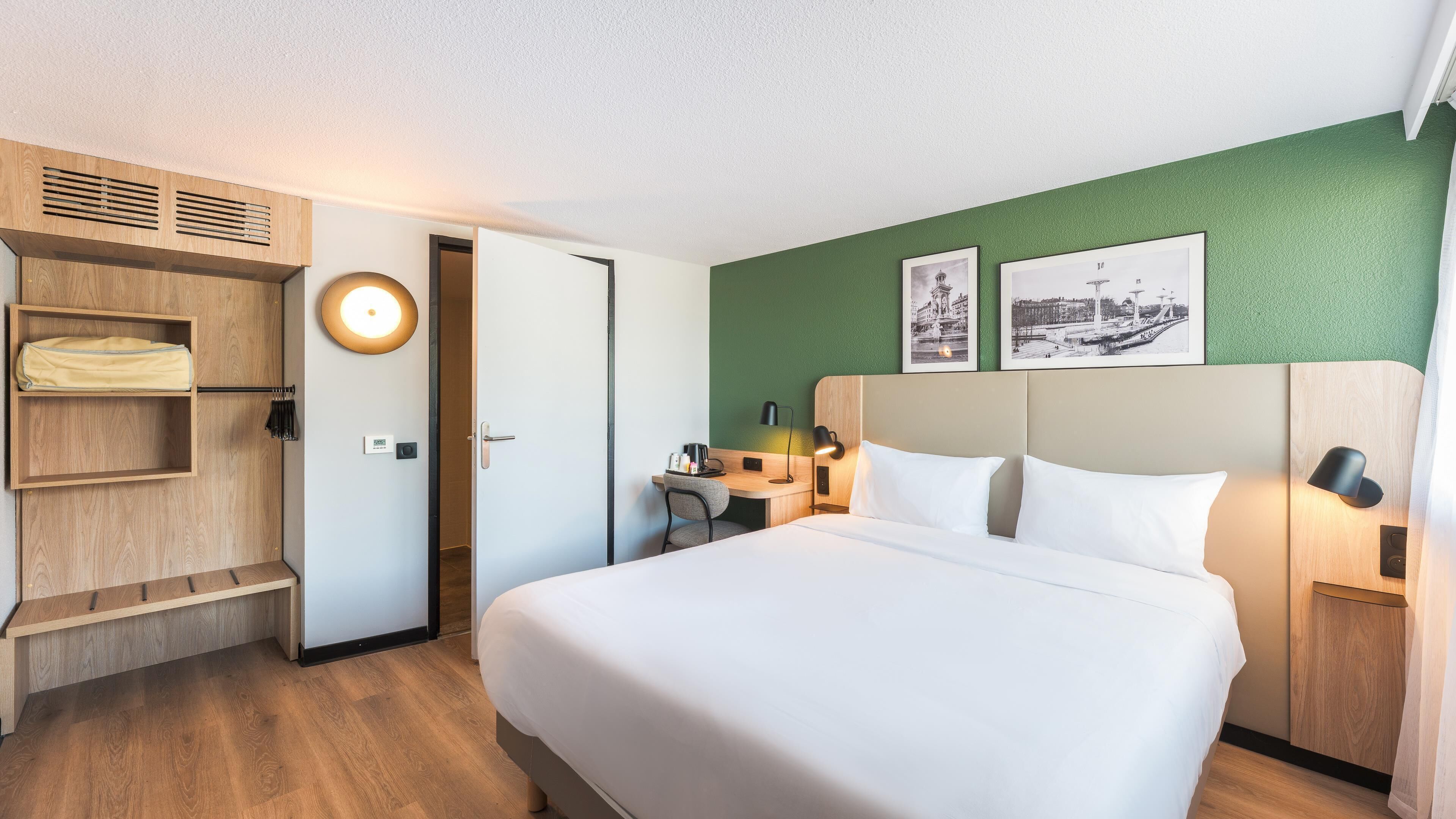 Habitación de hotel con cama, escritorio, silla, armario y dos lámparas fijadas a la pared.