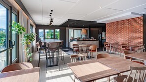 A modern dining area with a brick wall, large windows, and a central island with seating. - Campanile NATURE - Lyon Est Aéroport Saint Exupéry (Saint-Laurent-de-Mure)