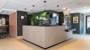 A modern reception area with a striped counter, a television, and a 'Self Check In' sign. - Campanile NATURE - Lyon Est Aéroport Saint Exupéry (Saint-Laurent-de-Mure)