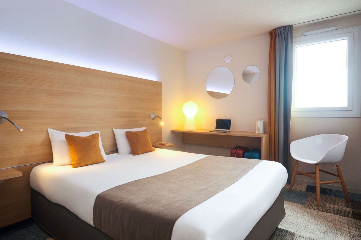 A hotel room with a large bed, a desk with a laptop, a chair, and a window with curtains.
