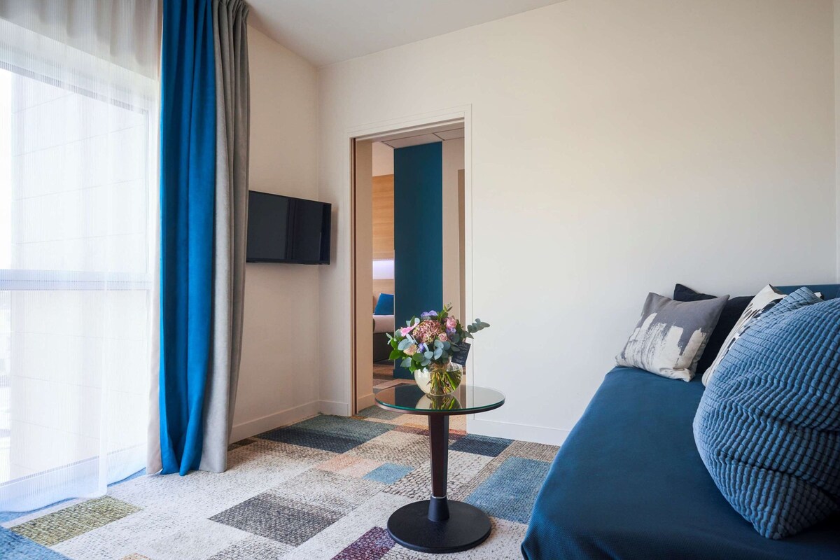 A hotel room with a bed, a glass table with a vase of flowers, a television on the wall, and a view of the hallway.
