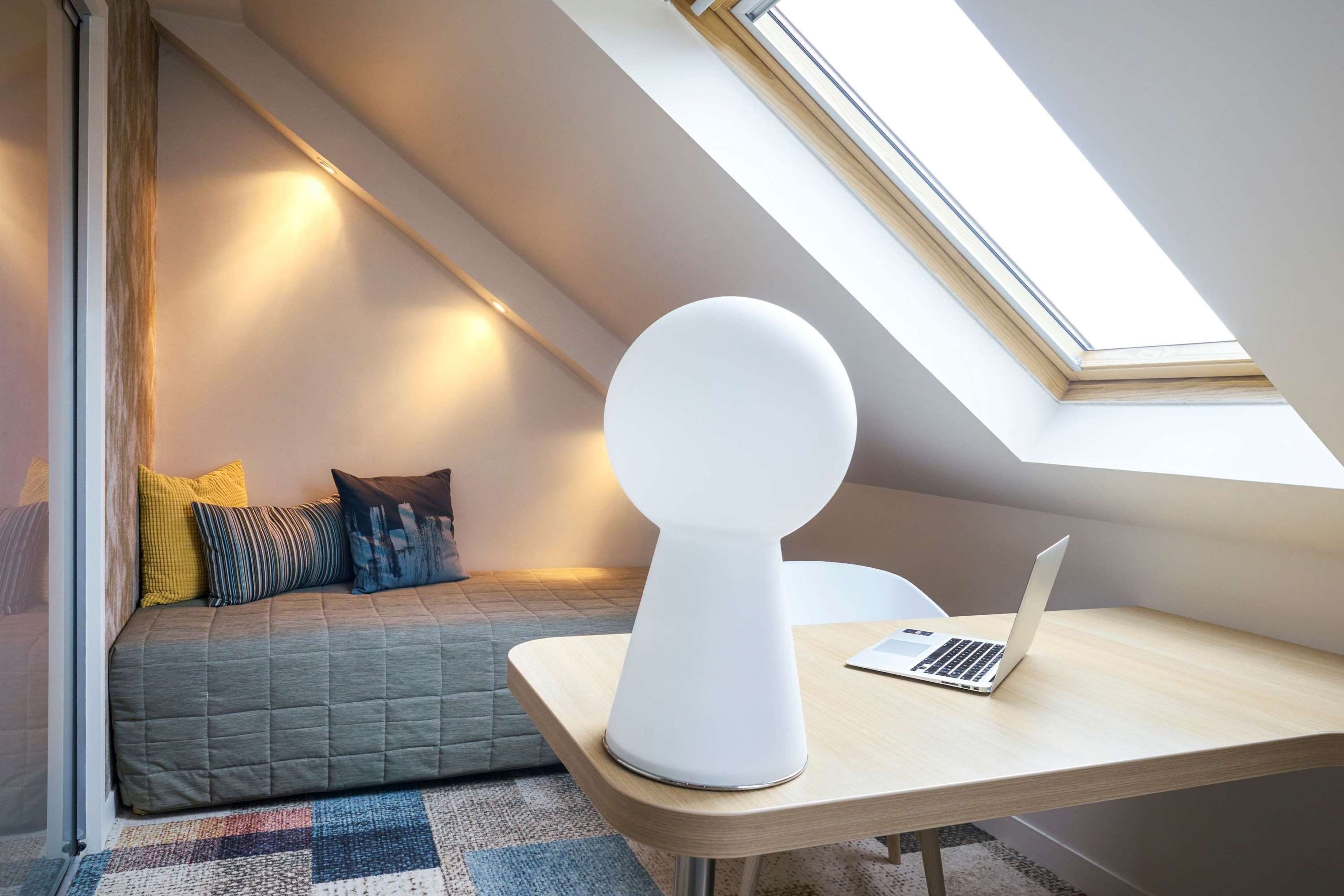 Quarto de loft moderno com claraboia, uma cama com almofadas coloridas e um laptop sobre uma escrivaninha de madeira.