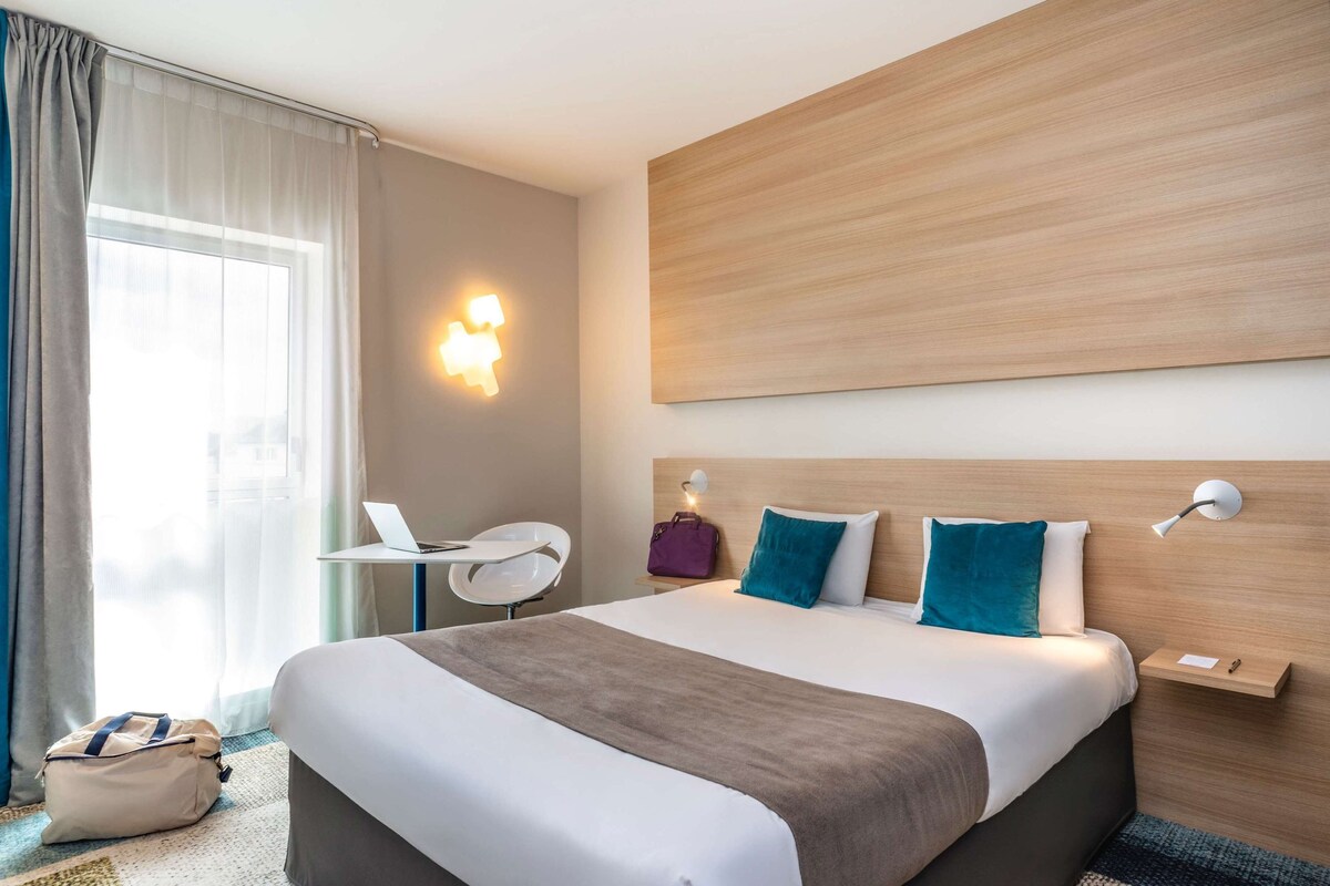 A hotel room with a bed, bedside table, lamp, and a view of the city through the window.