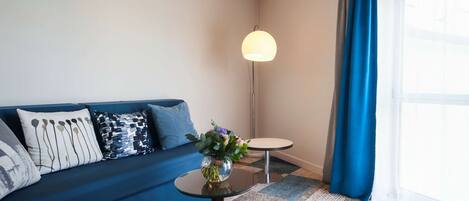 A modern living room with a blue sofa, a small round table with a vase of flowers, a floor lamp, and a checkered rug.