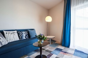 Ett modernt vardagsrum med en blå soffa, ett litet runt bord med en vas med blommor, en golvlampa och en rutig matta.