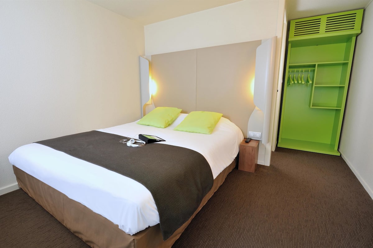 Un lit bien fait, recouvert d’une couvre-lit blanche et marron, avec deux oreillers verts et une table de chevet sur laquelle se trouvent une lampe et un téléphone.