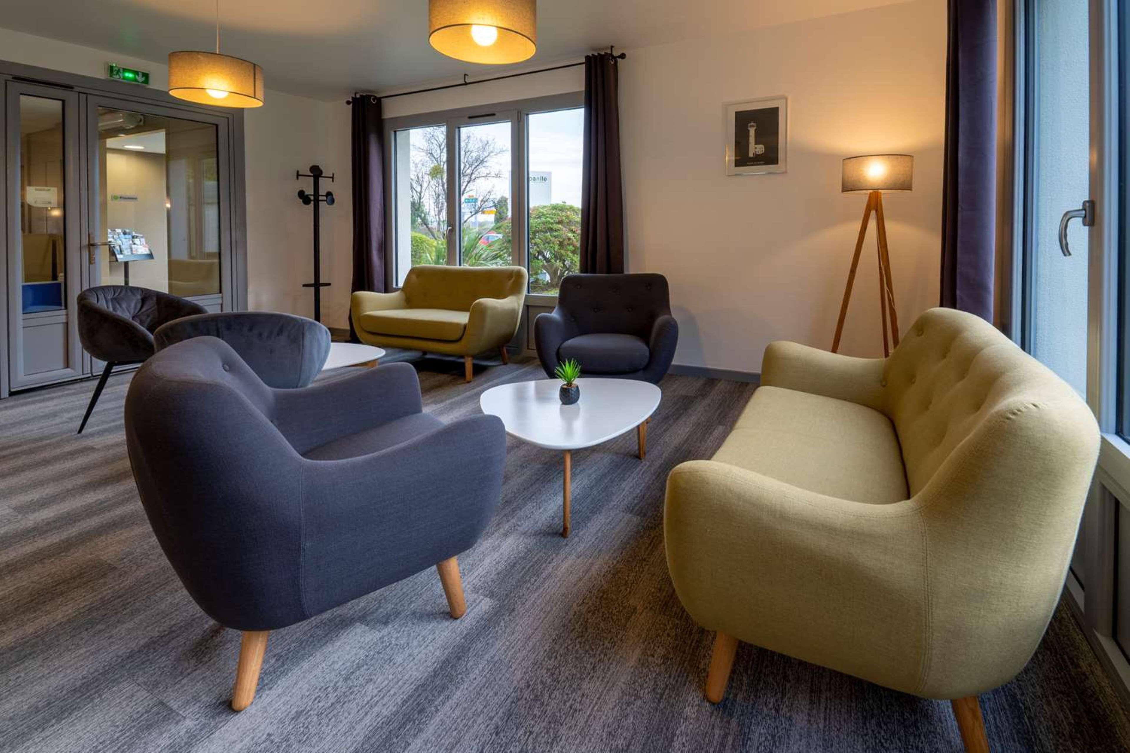 Habitación de hotel moderna con alfombra gris, dos sillones, una mesita redonda y una lámpara de pie.