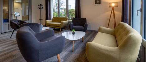 Quarto de hotel moderno com carpete cinza, duas poltronas, uma mesa redonda pequena e um abajur.