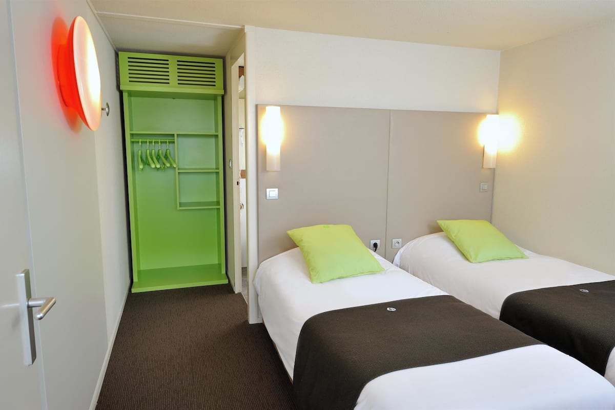 Une chambre d’hôtel avec deux lits, chacun équipé d’un oreiller, d’une armoire verte et d’un luminaire rouge.