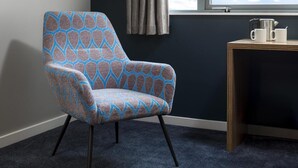 Una silla con estampado geométrico, una ventana con cortinas, una mesita con dos tazas blancas y un hervidor.