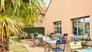 An outdoor patio with tables and chairs, a palm tree, and a building with large windows.