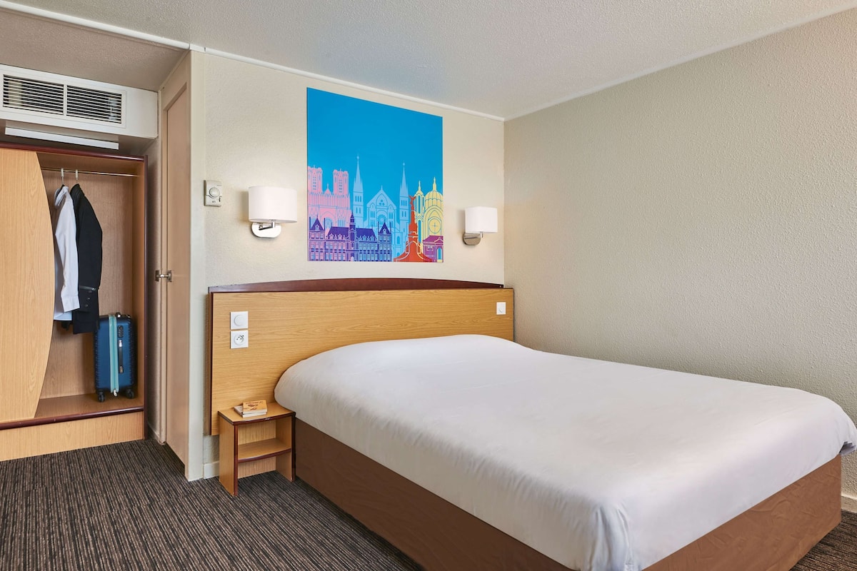 A hotel room with a bed, a wardrobe, a coat rack, a wall-mounted air conditioner, and a cityscape painting.