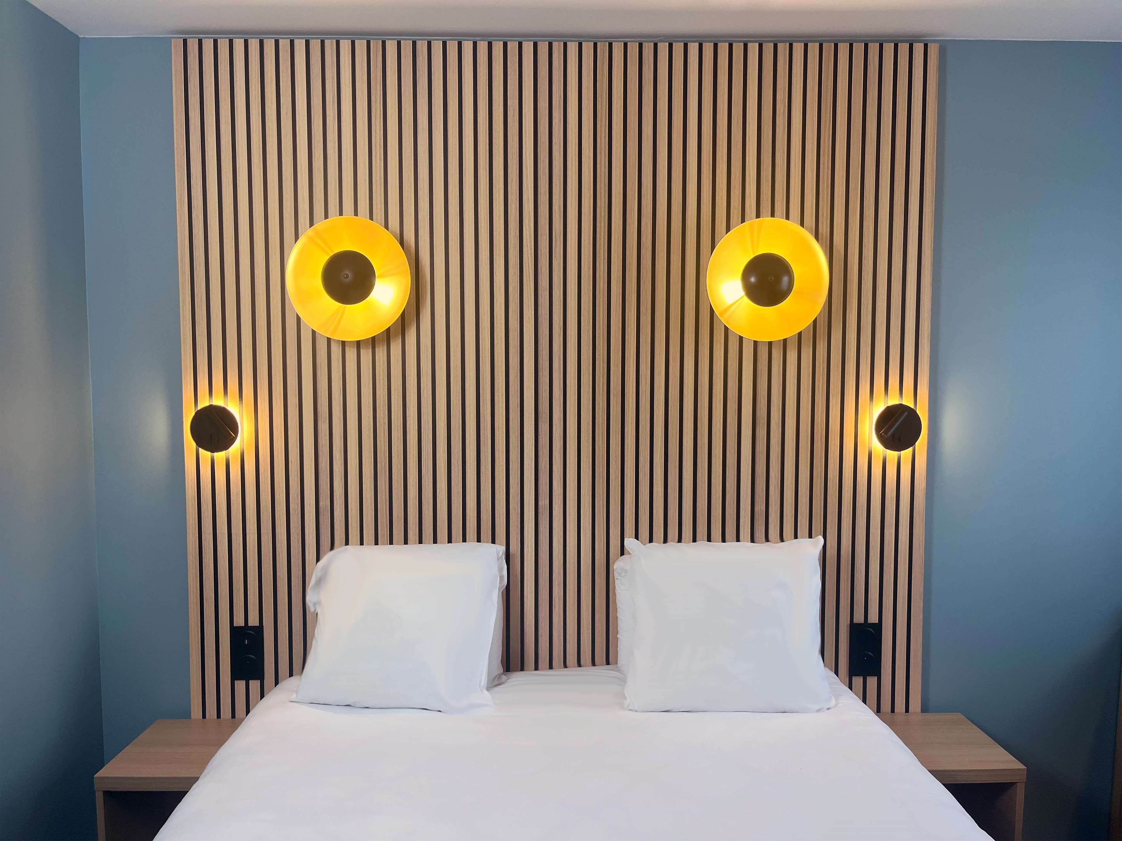 Ein Hotelzimmer mit einem Bett, zwei Nachttischen und einer Wand mit vertikalen Holzlatten und runden Wandleuchten.