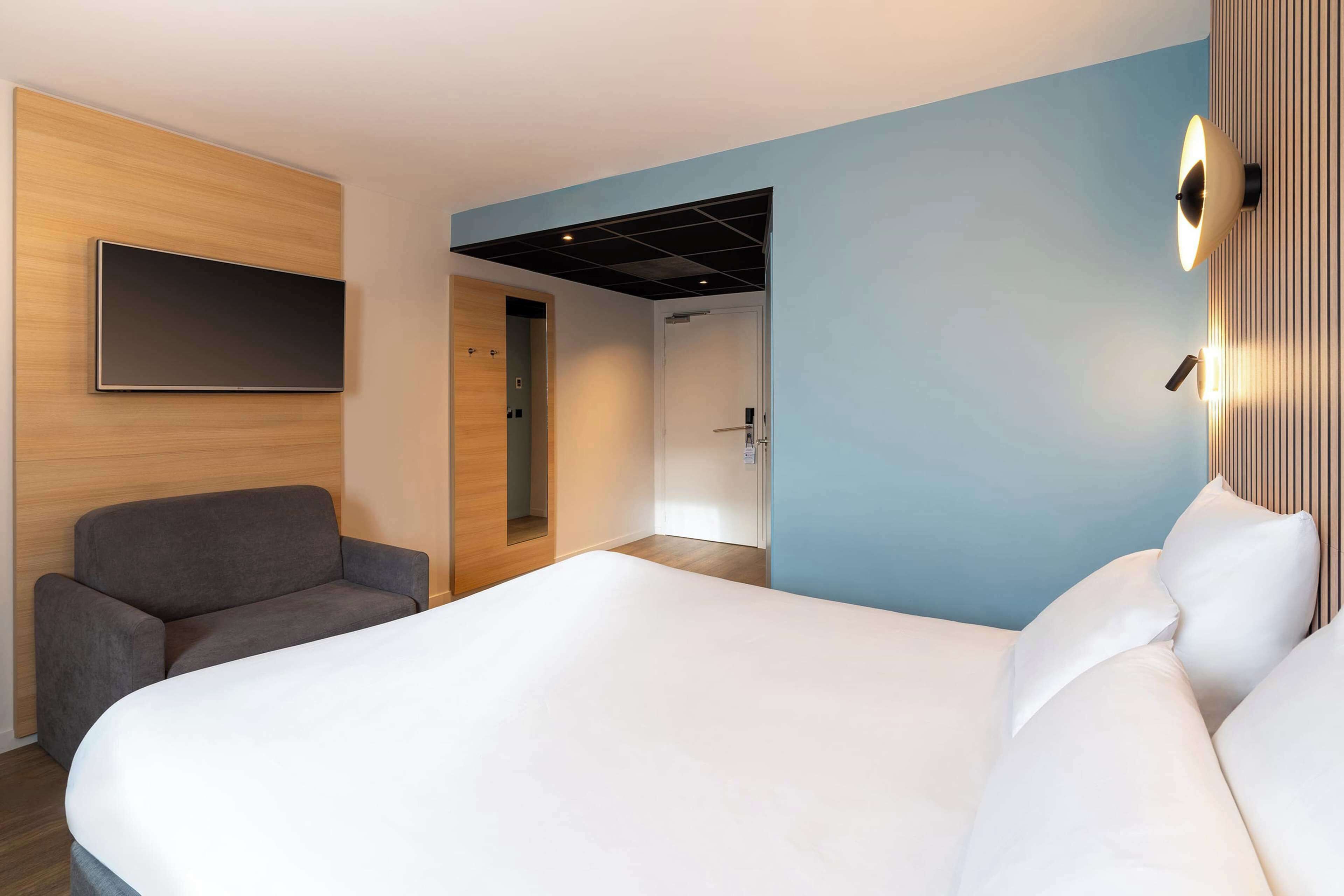 Una habitación de hotel moderna con una cama grande, un sofá gris, un televisor de pantalla plana montado en un panel de madera y una lámpara de pared.