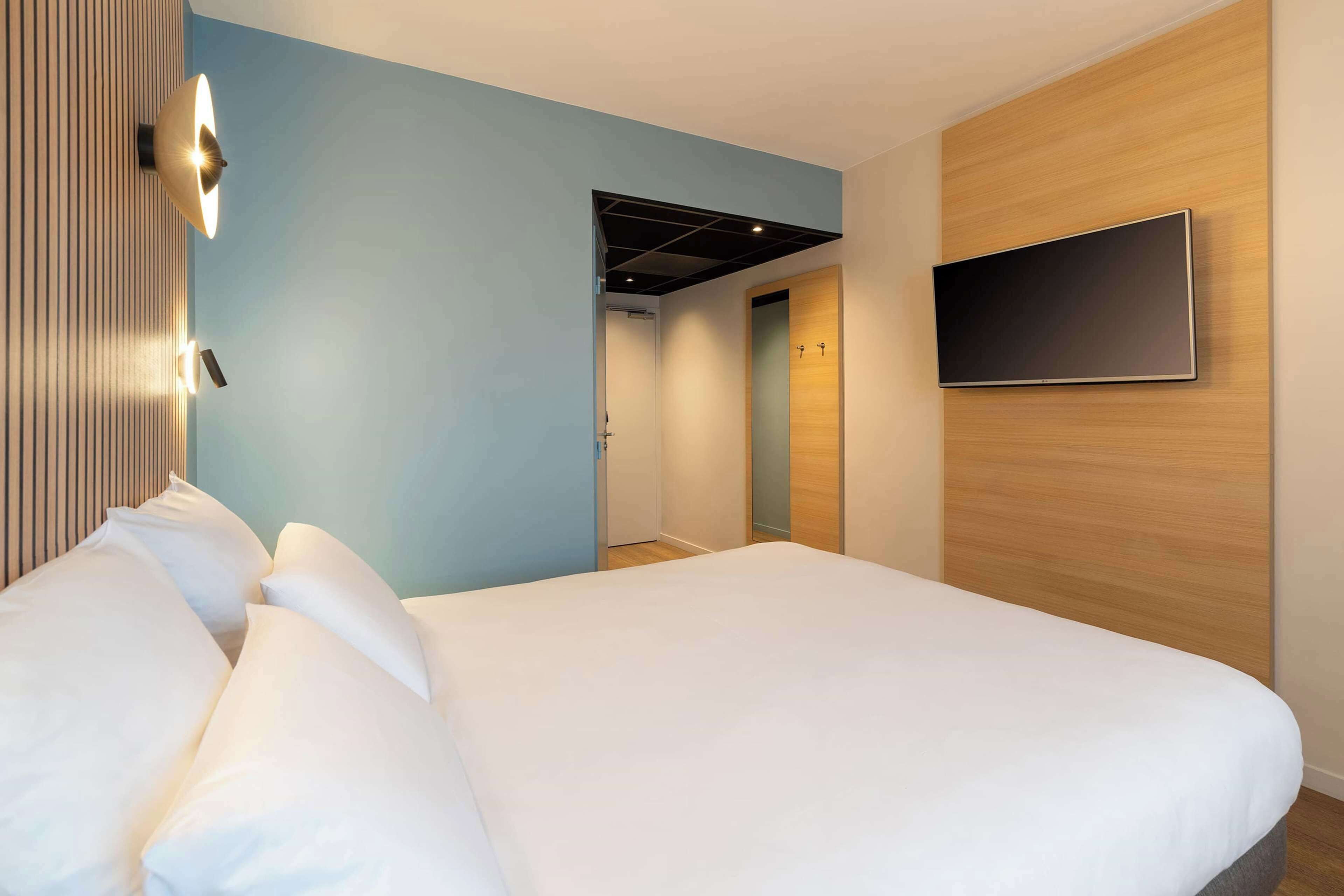 A modern hotel room with a large bed, a flat-screen TV mounted on the wall, and a wooden accent wall.