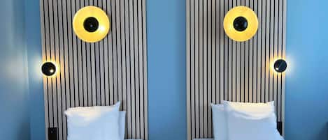 Habitación de hotel con dos camas, cada una con colchón y almohada blancos, flanqueadas por listones de madera verticales y luminarias circulares.