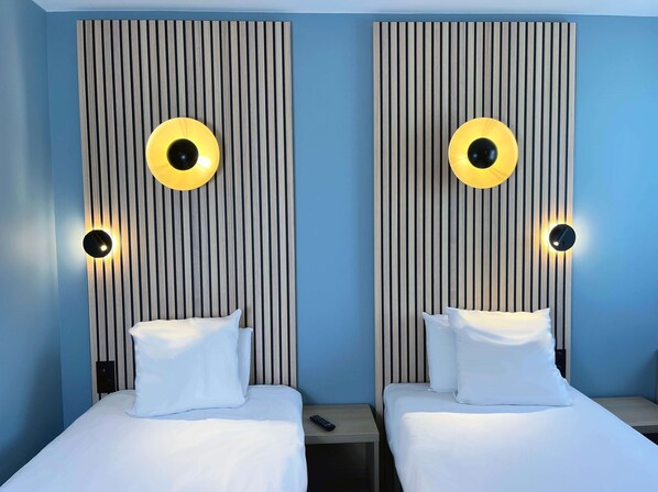 A hotel room with two beds, each with a white mattress and pillow, flanked by vertical wooden slats with circular light fixtures.