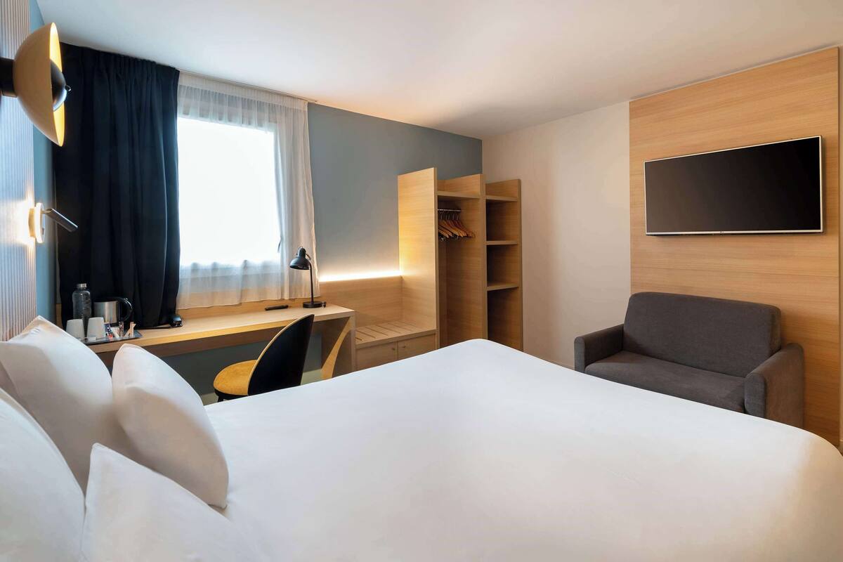 A hotel room with a large bed, a desk with a chair, a wardrobe, and a TV mounted on the wall.
