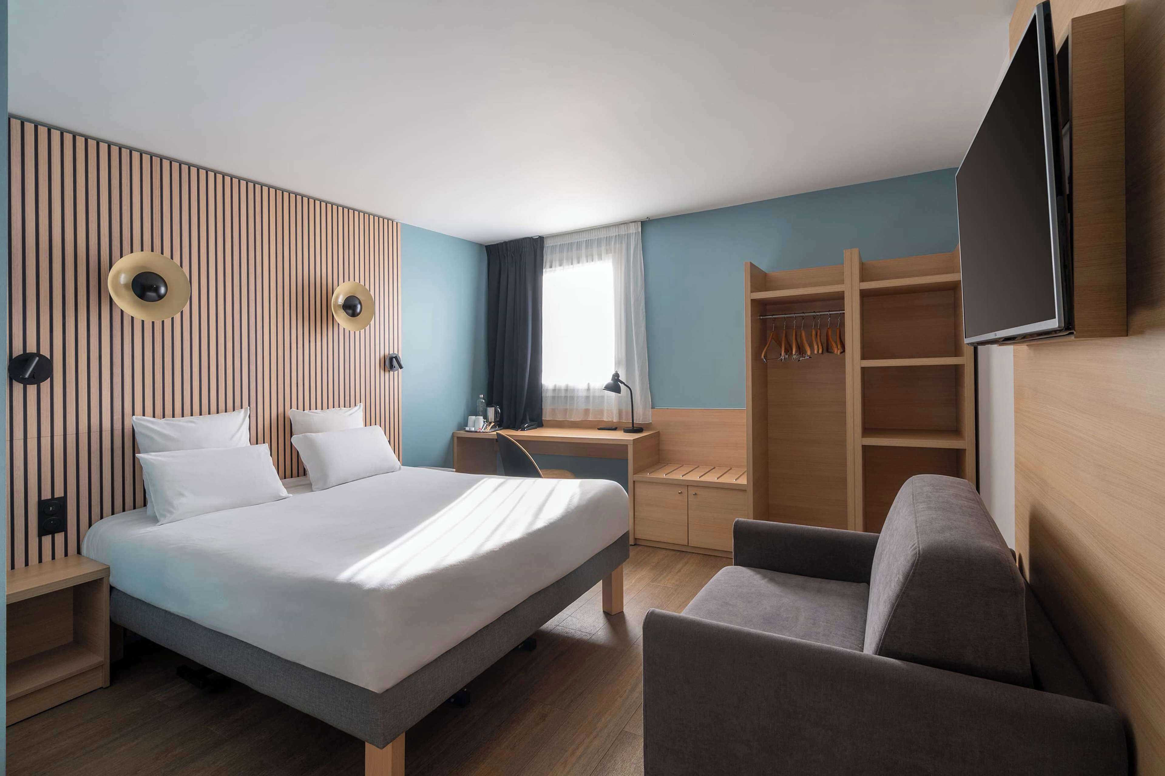 Camera d'albergo con letto, comodino, scrivania, sedia, televisore, armadio e finestra con tende.