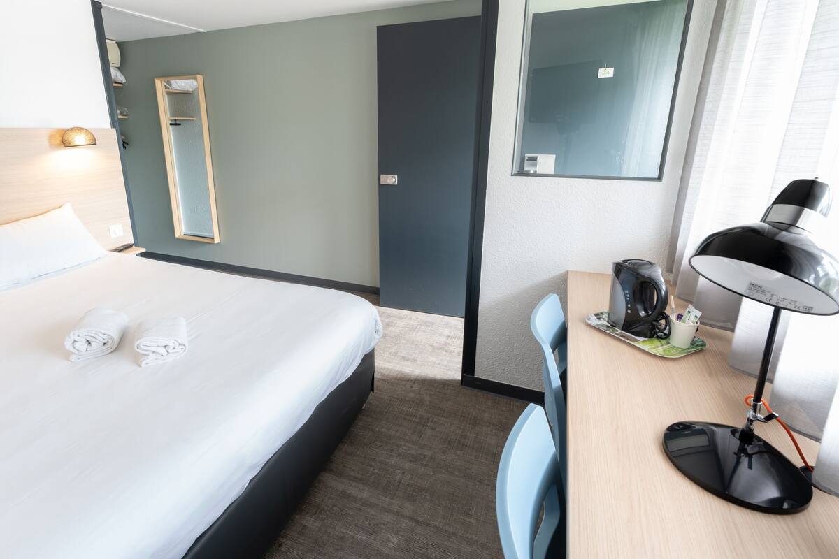 A hotel room with a bed, a desk, a chair, a mirror, and a coffee maker.