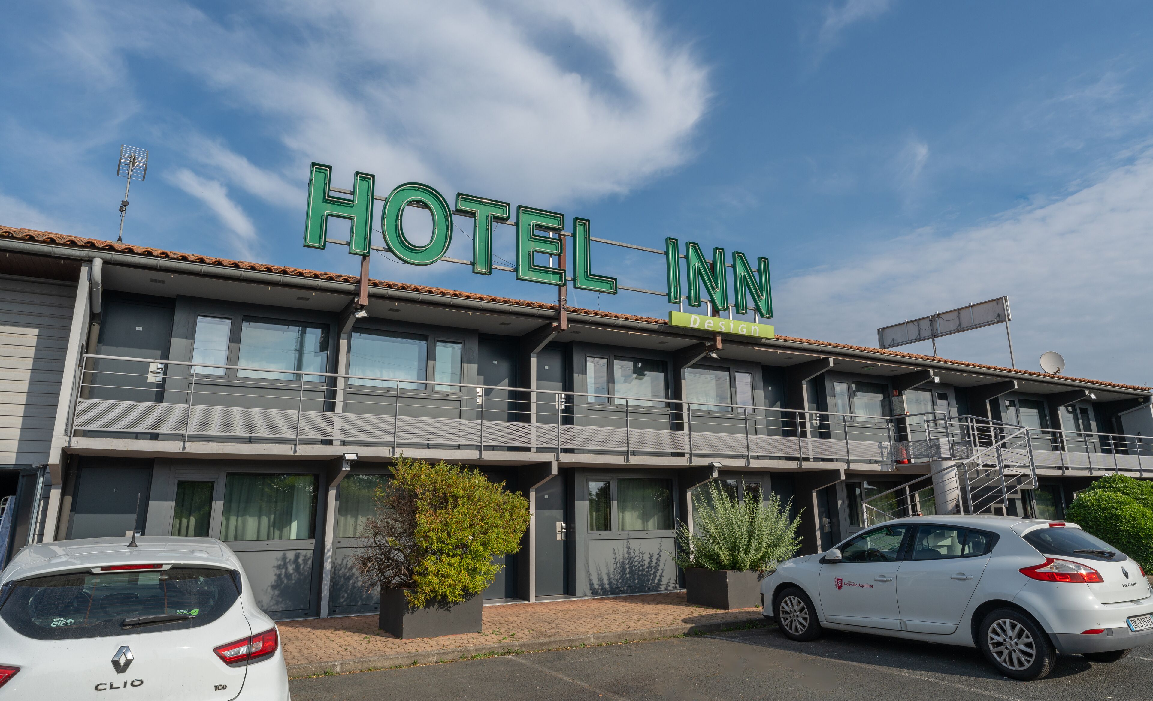 Un hôtel appelé « HOTEL INN », doté d’un design moderne, avec un balcon et une Renault Clio garée devant.