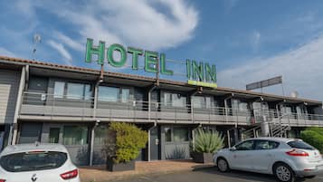 Un hotel llamado "HOTEL INN" con diseño moderno, que cuenta con un balcón y un automóvil Renault Clio estacionado frente a él.