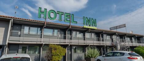 Un hôtel appelé « HOTEL INN », doté d’un design moderne, avec un balcon et une Renault Clio garée devant.