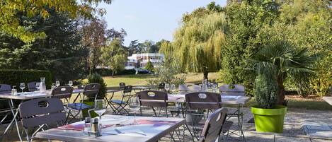 An outdoor dining area with tables and chairs, surrounded by trees and a building in the background.