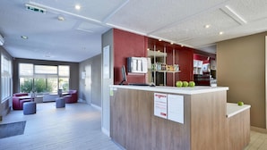 A modern reception area with a wooden counter, a seating area with red sofas, and a view of the outdoors through large windows. - Campanile Lyon Ecully (Ecully)