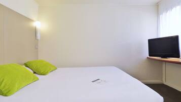 Hotellihuone, jossa on sänky, kaksi vihreää tyynyä, yöpöytä, jossa on televisio, ja seinälle asennettu valaisin.