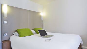 A neatly made bed with a laptop and a brochure, two green pillows, and a bedside table with a power outlet.
