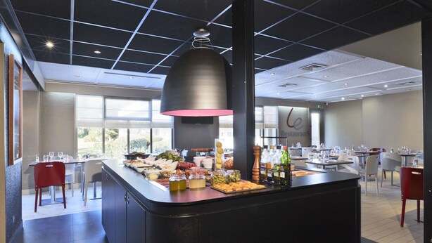 A modern restaurant with a central island featuring a variety of food and drink options, large windows, and a contemporary ceiling design. - Campanile Lyon Ecully (Ecully)