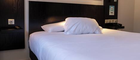A hotel room with a large bed, a headboard, a nightstand, a wall-mounted lamp, a framed cityscape picture, and a wall-mounted clock.