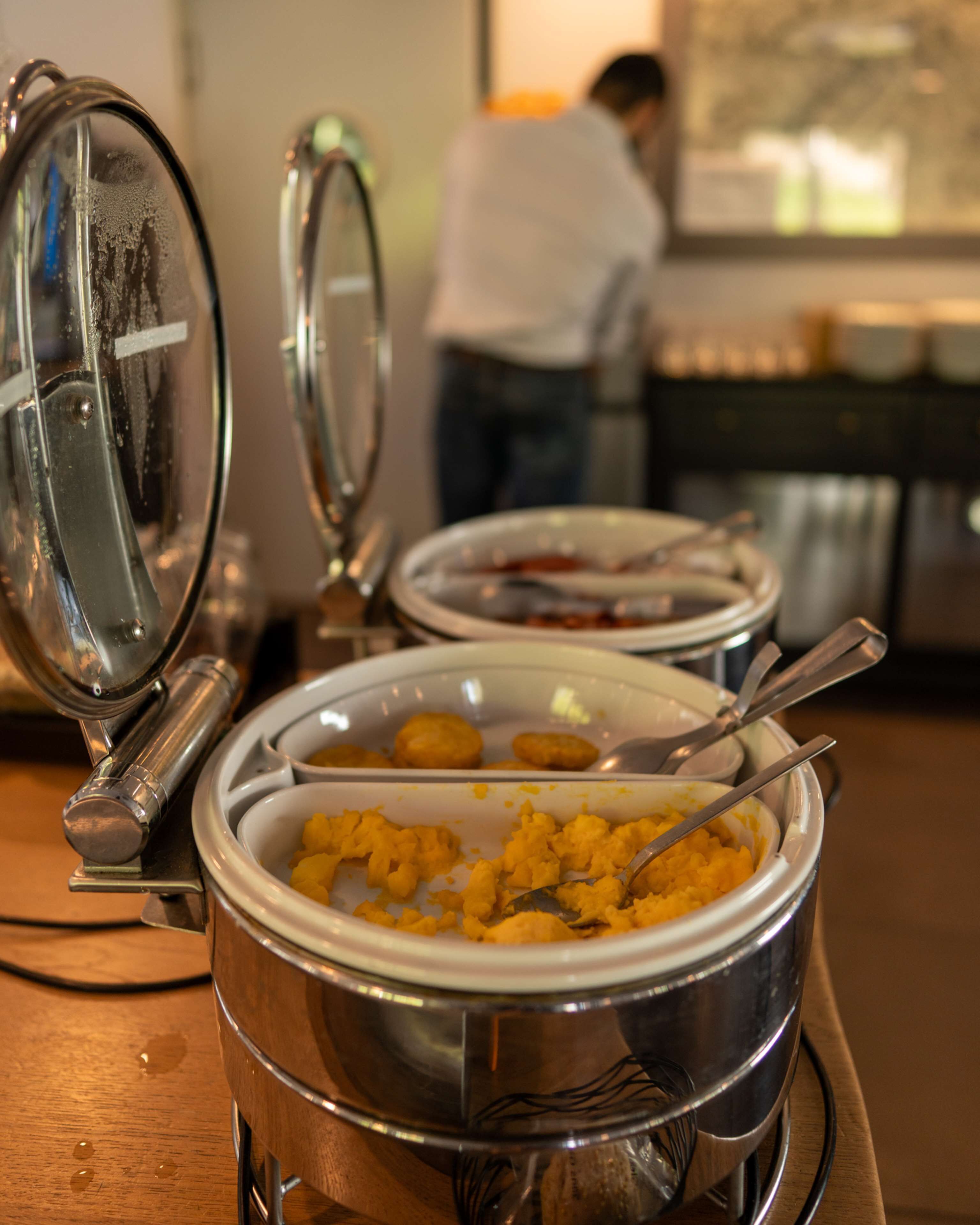 A buffet setup with various food items in metal serving dishes, a spoon in one dish, and a person in the background.