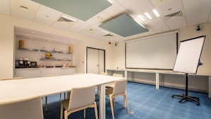 A conference room with a long table, chairs, a projector screen, and a whiteboard. - Mandarina Luxembourg Airport Hotel (Niederanven)