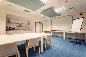 A conference room with a long table, chairs, a projector screen, and a whiteboard. - Mandarina Luxembourg Airport Hotel (Niederanven)