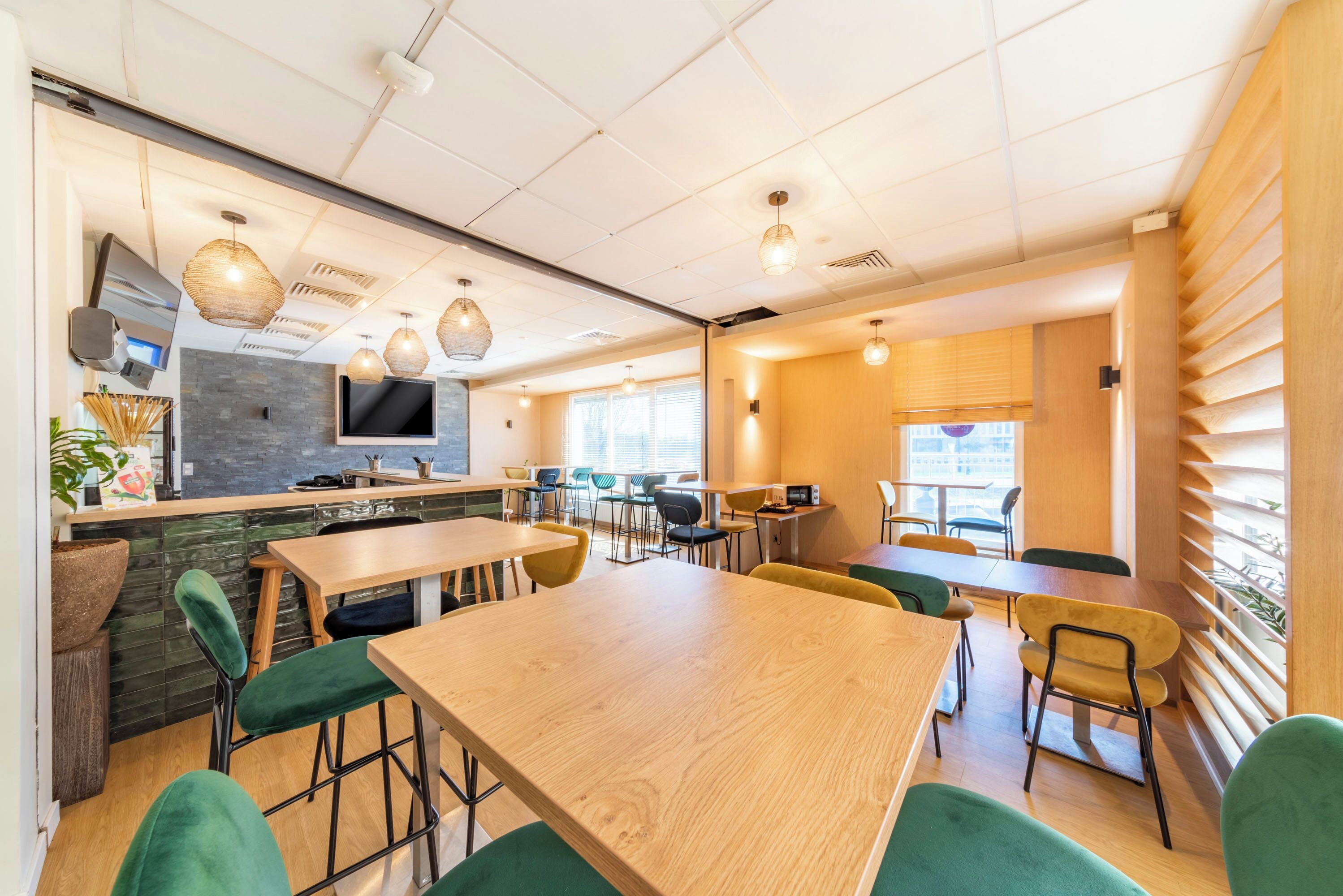 Un intérieur de restaurant moderne avec une longue table en bois, des chaises à revêtement vert et des luminaires suspendus.