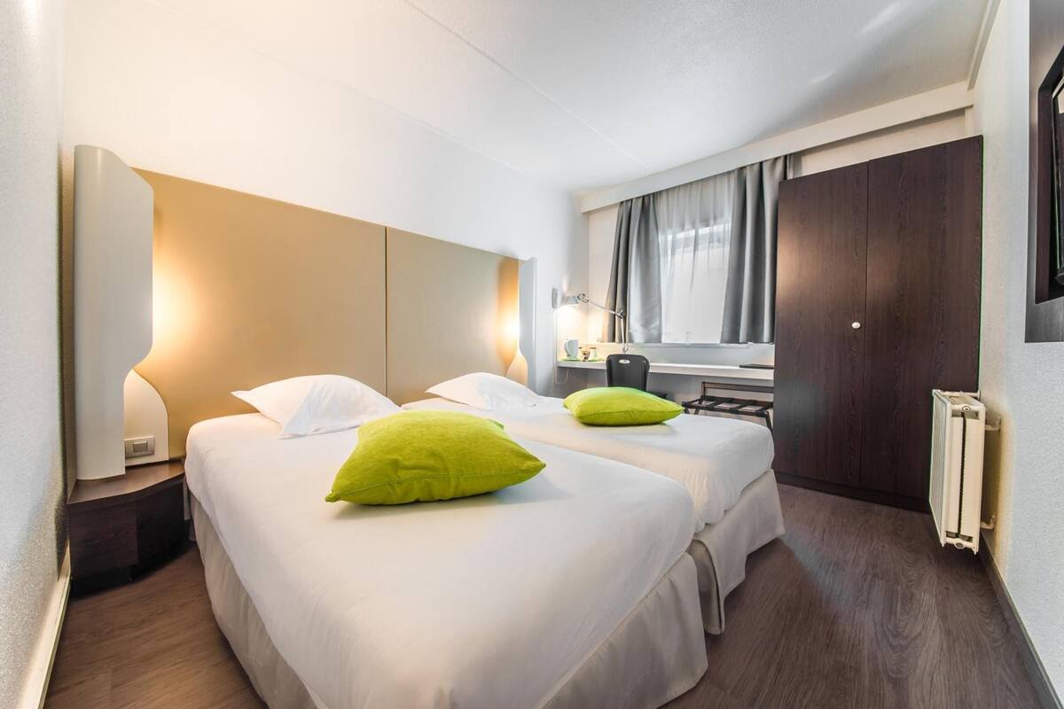 A hotel room with two beds, a desk, a chair, a window with curtains, and a radiator.