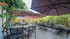 An outdoor patio area with multiple tables and chairs, shaded by large umbrellas, surrounded by greenery. - Mandarina Luxembourg Airport Hotel (Niederanven)