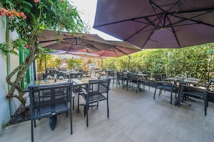 An outdoor patio area with multiple tables and chairs, shaded by large umbrellas, surrounded by greenery.
