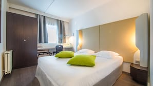 A hotel room with a large bed, two green pillows, a desk with a chair, a radiator, and a nightstand with a lamp. - Mandarina Luxembourg Airport Hotel (Niederanven)
