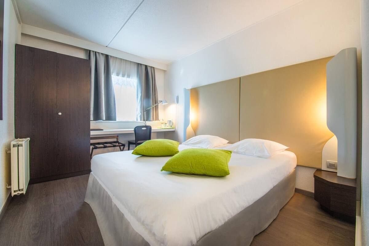 A hotel room with a large bed, two green pillows, a desk with a chair, a radiator, and a nightstand with a lamp.