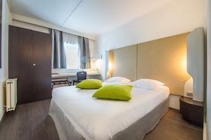 A hotel room with a large bed, two green pillows, a desk with a chair, a radiator, and a nightstand with a lamp.