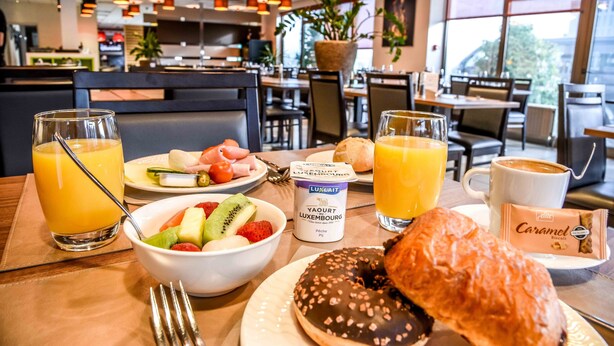 A breakfast setting with a fruit bowl, croissant, juice, coffee, and a pastry on a table. - Mandarina Luxembourg Airport Hotel (Niederanven)