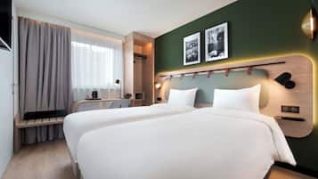 A hotel room with two beds, a desk, a chair, a window with curtains, and two framed pictures on the wall.
