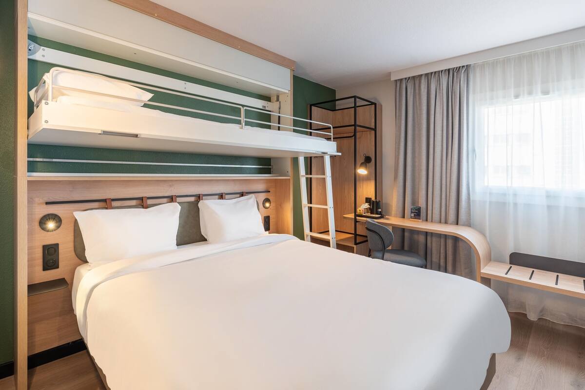 A hotel room with a bunk bed, a desk with a chair, a small table, and a window with curtains.